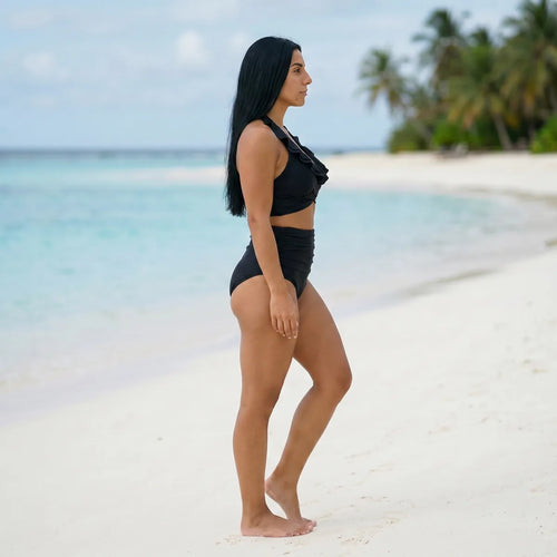 Bikini taille haute gainant noir Elya — silhouette galbée vue de profil sur plage paradisiaque